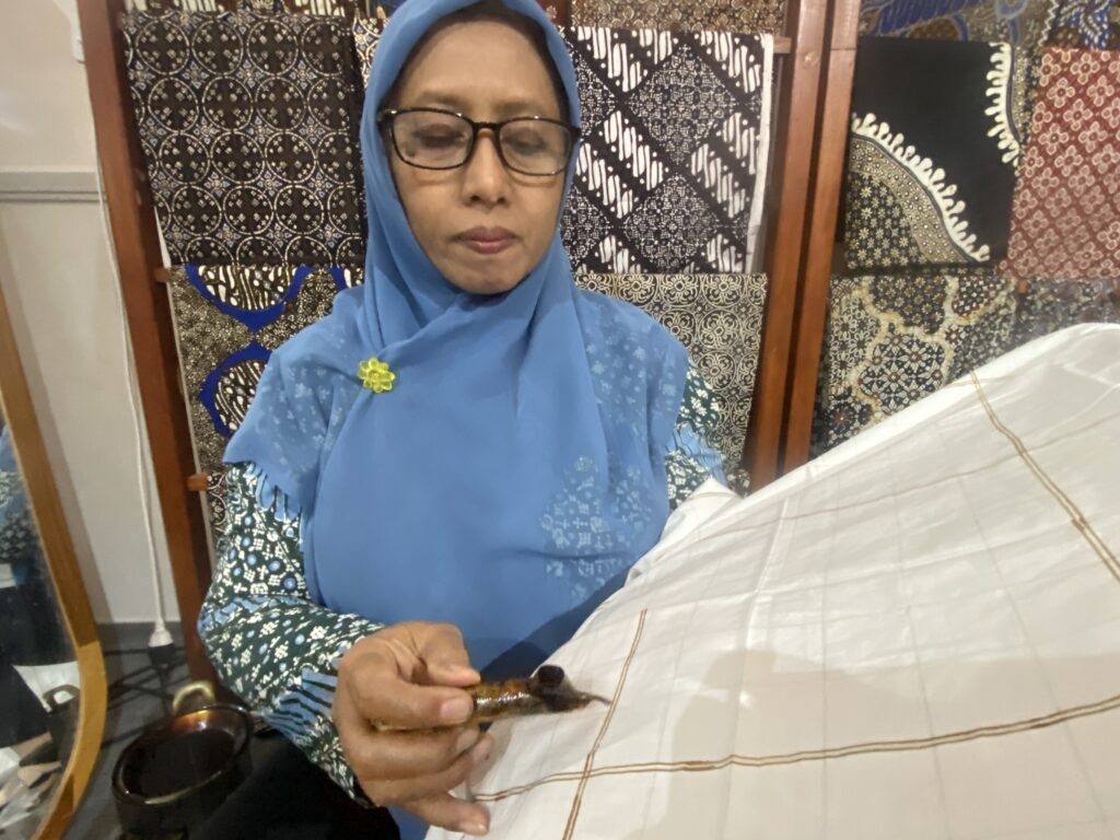 Woman wearing a blue hijab and glasses sits at a table, holding a small brown glass bottle in her hands with patterned fabric behind her.