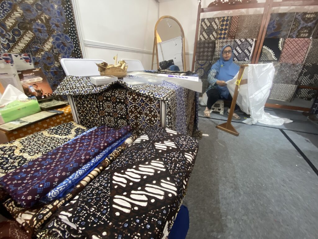 Textile shop interior with shelves and tables covered in patterned fabrics; a woman in a blue hijab sits nearby, looking at camera.