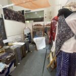 Fabric booth with patterned textiles on tables and racks; mannequin in a blue-and-white patterned dress on the right.