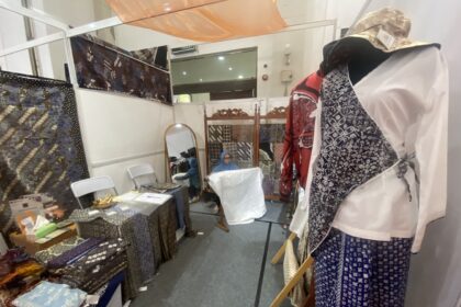 Fabric booth with patterned textiles on tables and racks; mannequin in a blue-and-white patterned dress on the right.