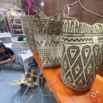 Rows of black-and-natural woven baskets on a wooden display, with a person weaving in the background at a market stall