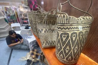Rows of black-and-natural woven baskets on a wooden display, with a person weaving in the background at a market stall