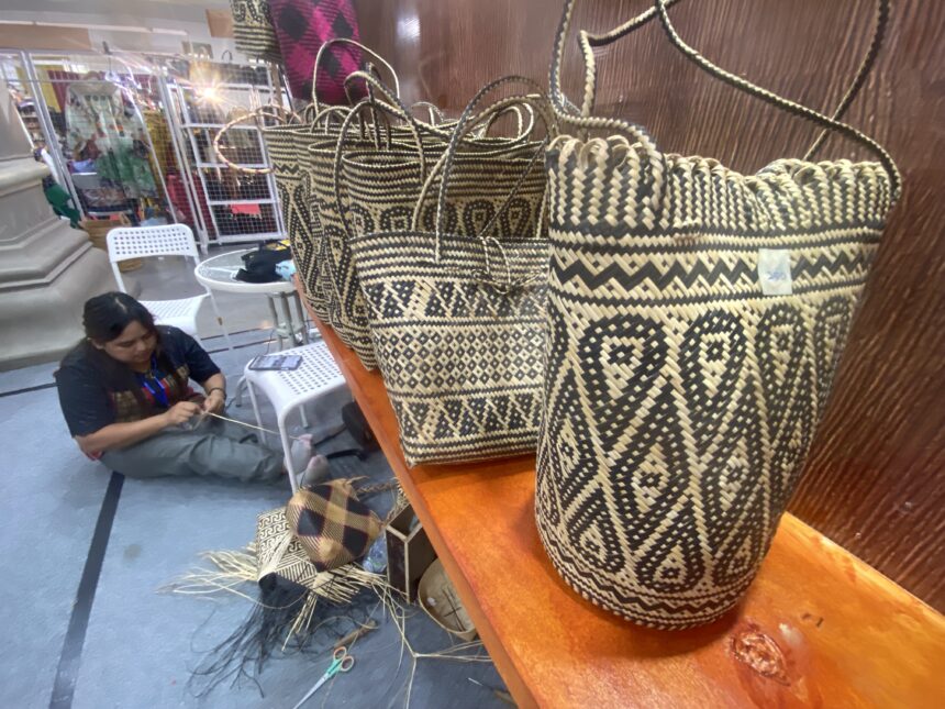 Rows of black-and-natural woven baskets on a wooden display, with a person weaving in the background at a market stall