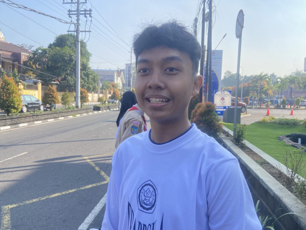 IMG_0515 - Mabur.co Young man smiling while taking a selfie on a sunny urban street, with a road, cars, and power lines in the background.