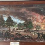 Battle scene in a tropical garden: soldiers fire, people flee, and a thatched pavilion burns in the background.