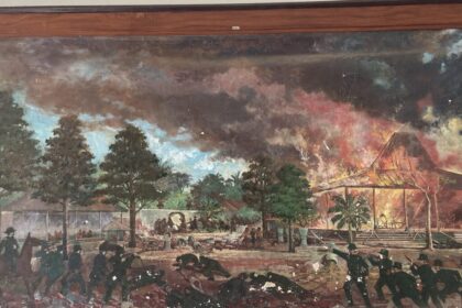 Battle scene in a tropical garden: soldiers fire, people flee, and a thatched pavilion burns in the background.