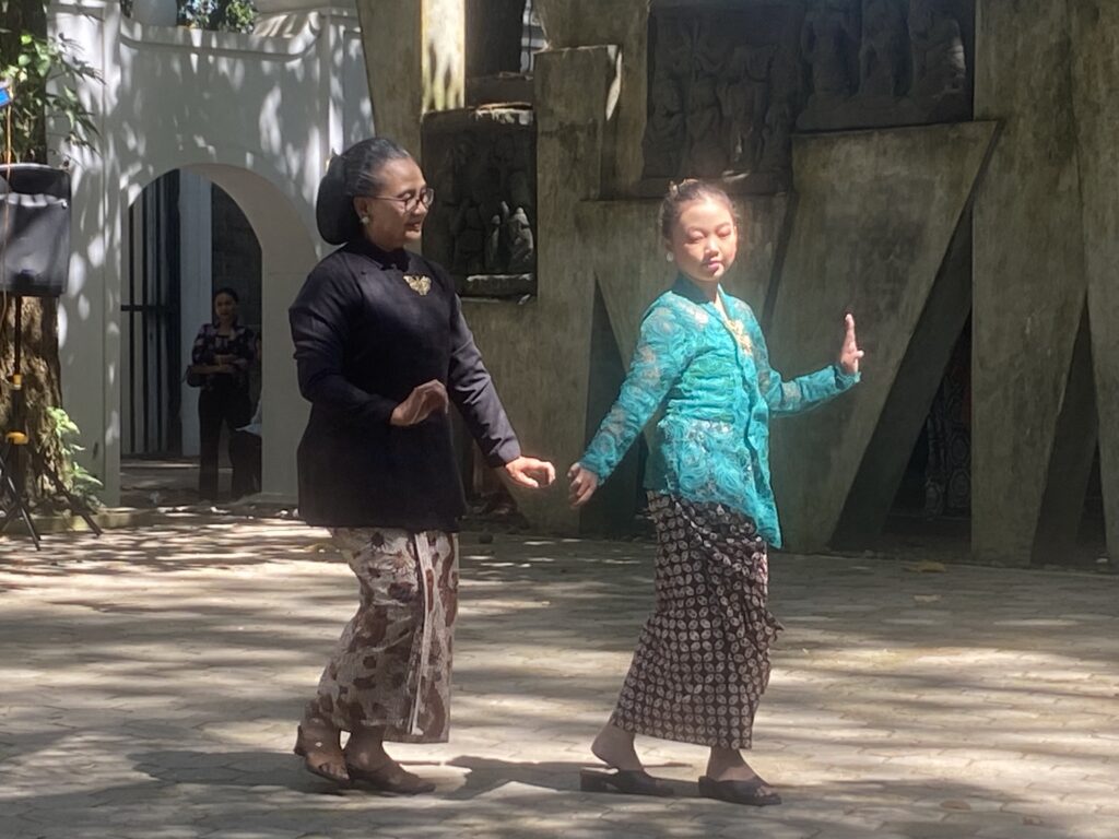 Two women dance outdoors, hand in hand, wearing traditional batik skirts and colorful tops under dappled sunlight.