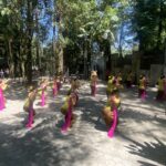 Women dancers in yellow outfits with pink sashes perform a choreographed routine outdoors among trees, with a fence and audience in the background.