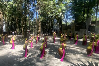 Women dancers in yellow outfits with pink sashes perform a choreographed routine outdoors among trees, with a fence and audience in the background.