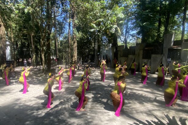 Women dancers in yellow outfits with pink sashes perform a choreographed routine outdoors among trees, with a fence and audience in the background.