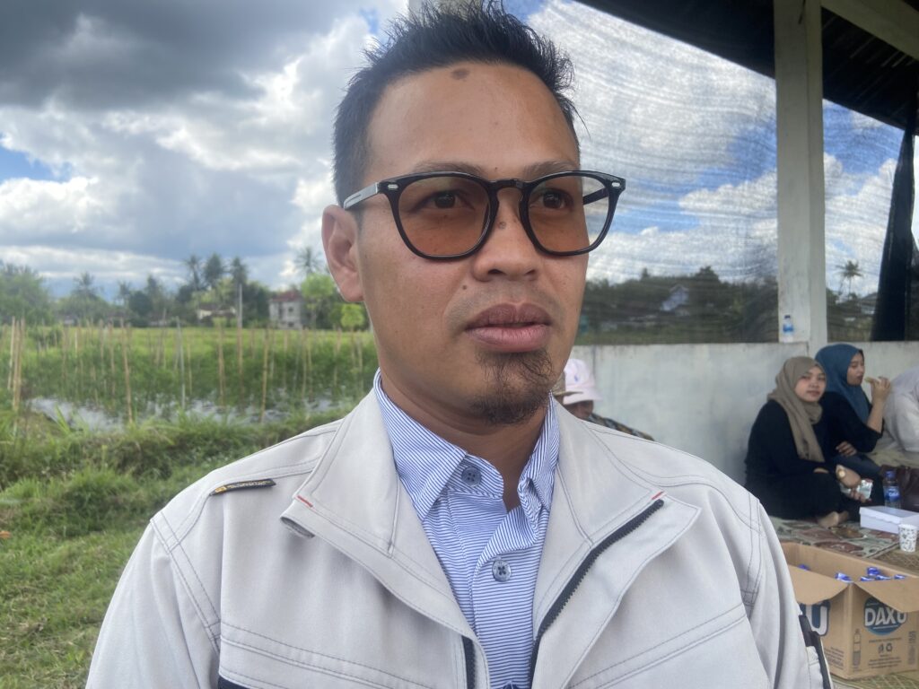 Close-up of a man in a light jacket wearing black-rimmed glasses, taking a selfie outdoors with a cloudy sky behind him. In the background, people sit by boxes under a shelter.