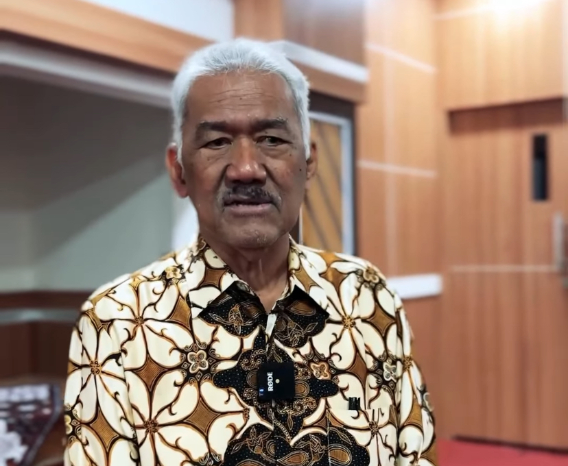 Portrait of an older man with white hair and mustache wearing a brown-and-cream batik shirt, indoors with wood-paneled walls.
