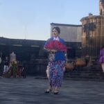 Woman in a blue and pink traditional dress holding a bright pink fan on an outdoor stage.