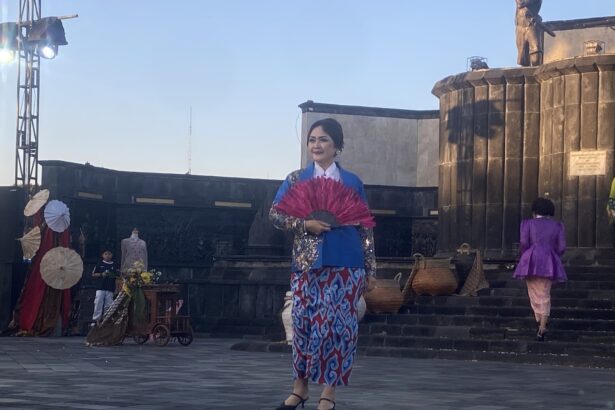 Woman in a blue and pink traditional dress holding a bright pink fan on an outdoor stage.