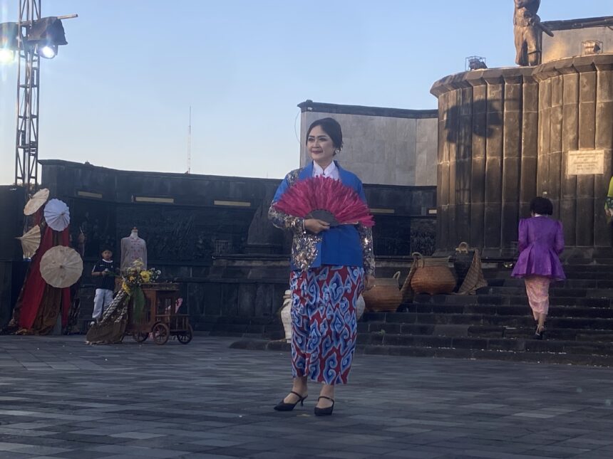 Woman in a blue and pink traditional dress holding a bright pink fan on an outdoor stage.