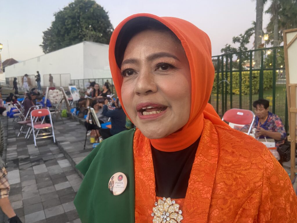 Close-up of a woman in an orange hijab smiling at an outdoor event, with people and chairs in the background.",