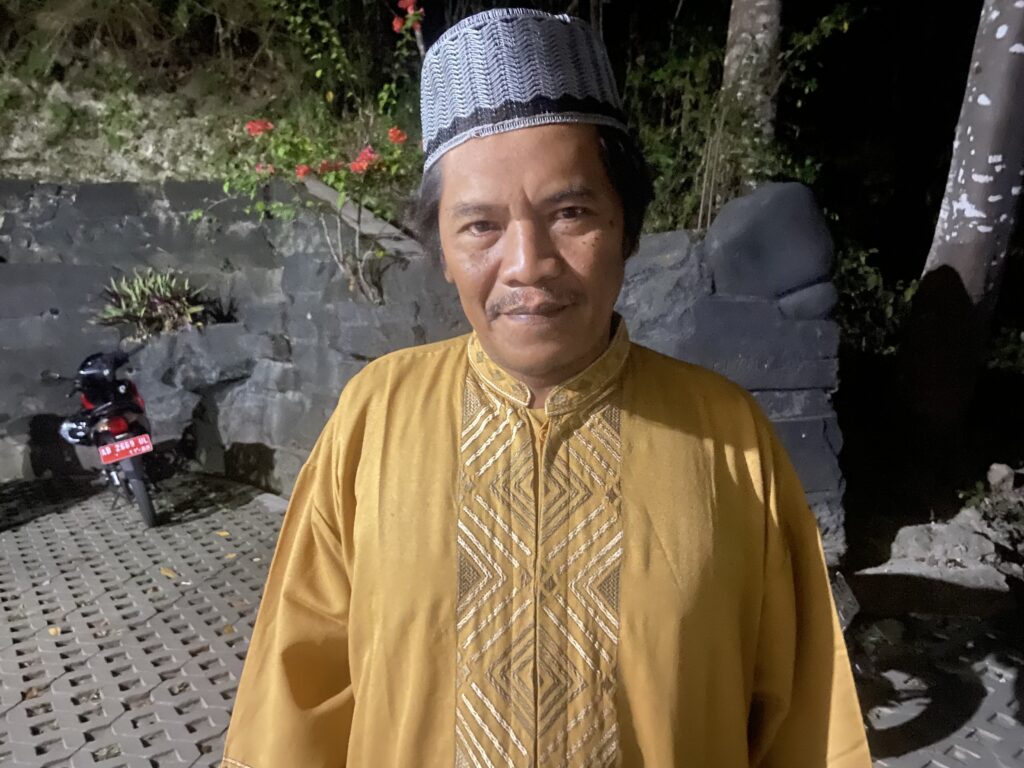 IMG_0857 - Mabur.co Man wearing a yellow traditional outfit with patterned sash, standing at night outdoors, smiling, with a stone wall, plants, and a motorcycle in the background.