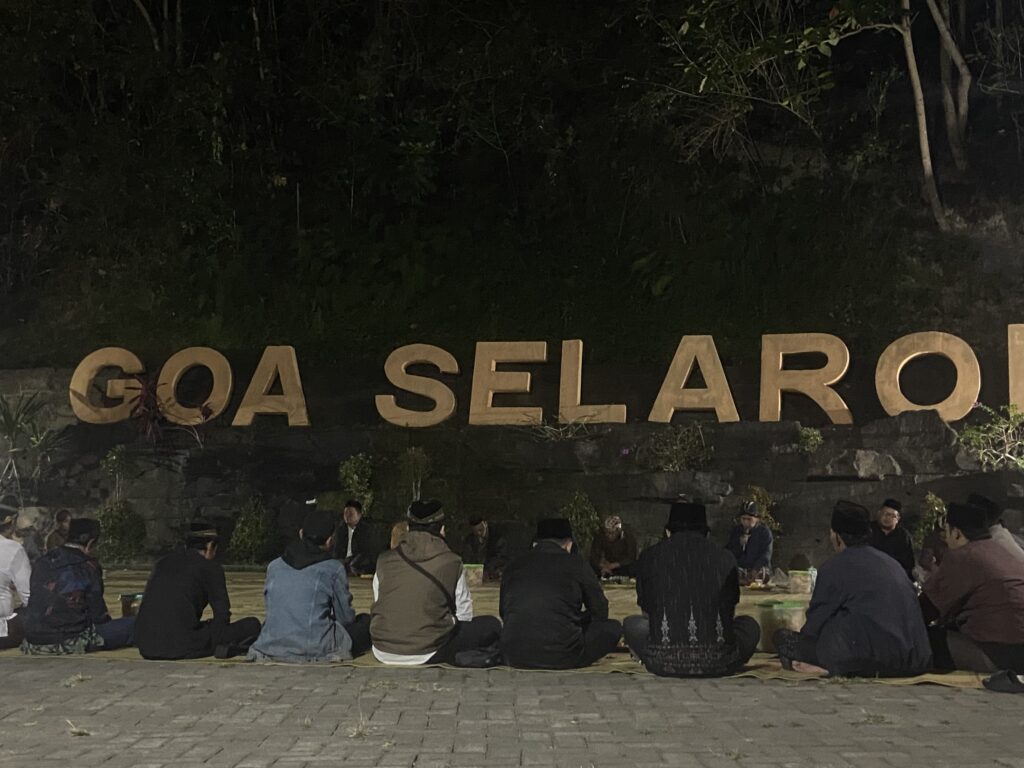 IMG_0863 - Mabur.co Group of people sitting cross-legged on a paved surface at night, arranged in rows with a large sign behind them.