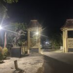Nighttime entrance with decorative gate, lit by street lamps and a 'Selamat Datang' sign in front of a building courtyard.