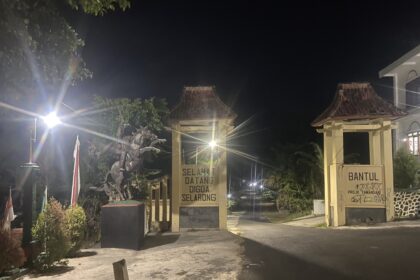 Nighttime entrance with decorative gate, lit by street lamps and a 'Selamat Datang' sign in front of a building courtyard.