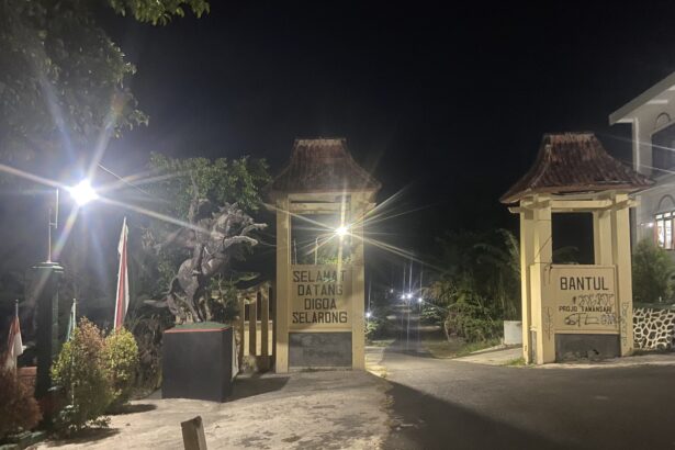 Nighttime entrance with decorative gate, lit by street lamps and a 'Selamat Datang' sign in front of a building courtyard.