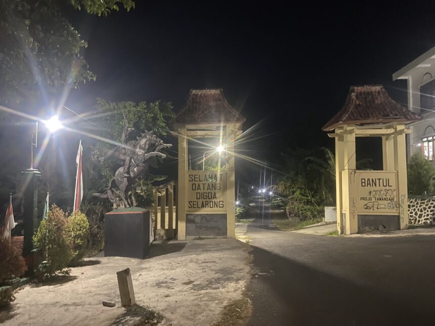 Nighttime entrance with decorative gate, lit by street lamps and a 'Selamat Datang' sign in front of a building courtyard.