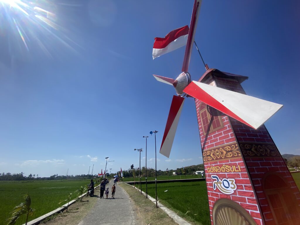 IMG_0903 - Mabur.co Red-and-white decorative windmill sculpture beside a walkway in a sunny park under a clear blue sky.