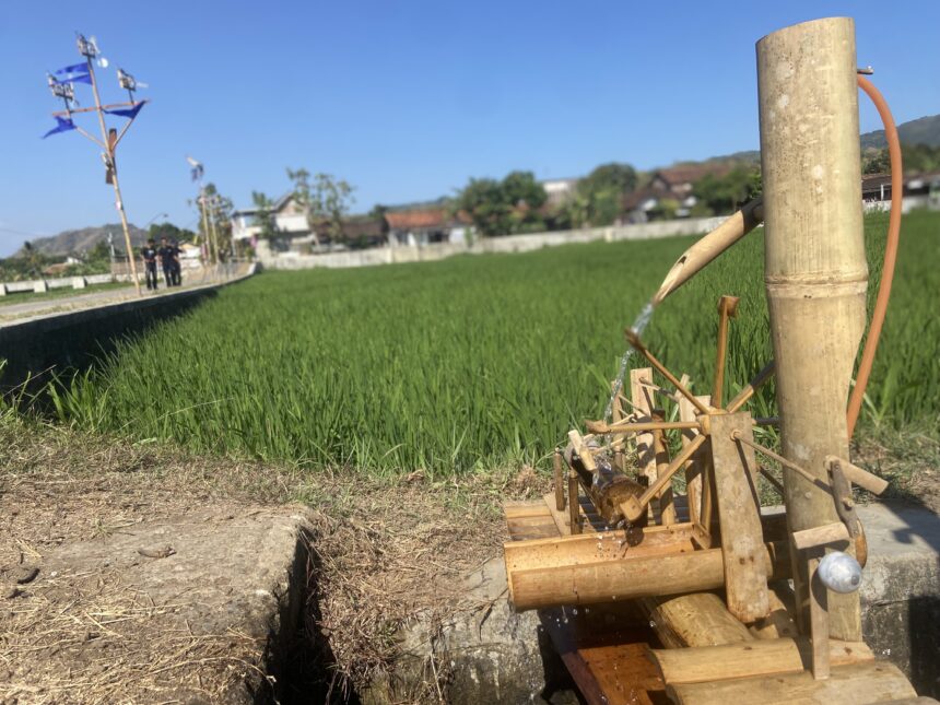 Close-up of a traditional bamboo water pump in a rice field, with a village and blue sky in the background.