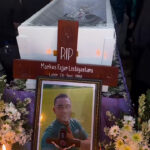 Wooden cross memorial with RIP and the name Markus Fajar Listiyantara on a coffin, surrounded by flowers.