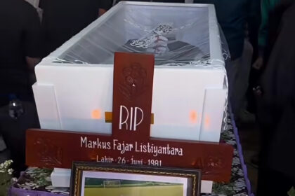 Wooden cross memorial with RIP and the name Markus Fajar Listiyantara on a coffin, surrounded by flowers.