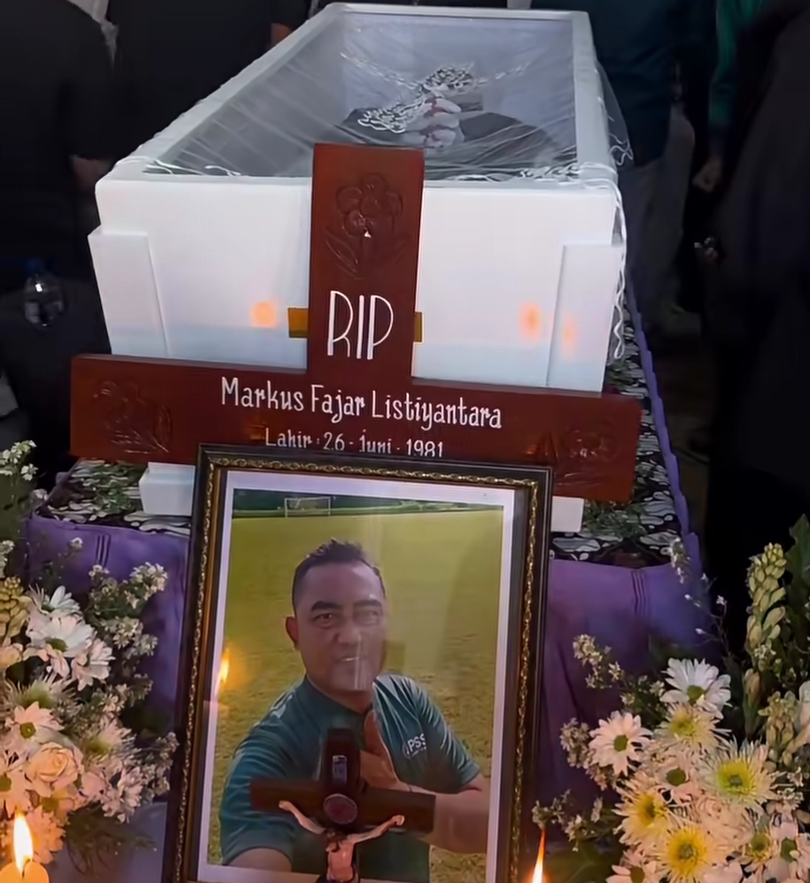 Wooden cross memorial with RIP and the name Markus Fajar Listiyantara on a coffin, surrounded by flowers.