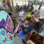 Group of people making paper crafts on mats under a pavilion at a daytime outdoor workshop.