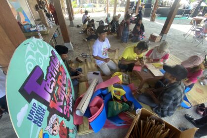 Group of people making paper crafts on mats under a pavilion at a daytime outdoor workshop.
