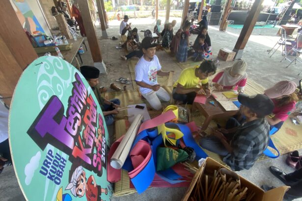 Group of people making paper crafts on mats under a pavilion at a daytime outdoor workshop.