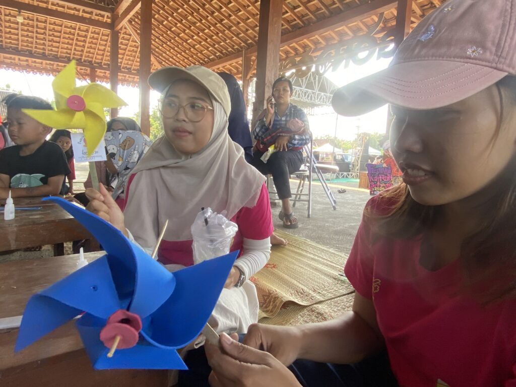 IMG_0965 1 - Mabur.co People gathered under a wooden pavilion doing a crafts activity; a woman in a hijab and glasses sits at the table with blue paper windmill crafts in progress.