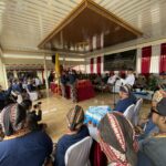 Ceremonial gathering in a hall with people in traditional Indonesian attire seated around a decorated stage and banner in the background