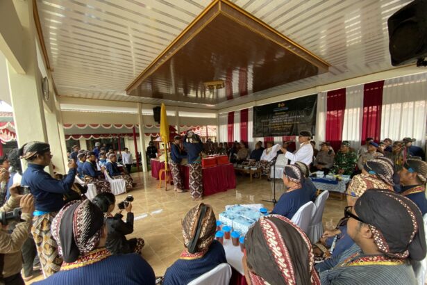 Ceremonial gathering in a hall with people in traditional Indonesian attire seated around a decorated stage and banner in the background