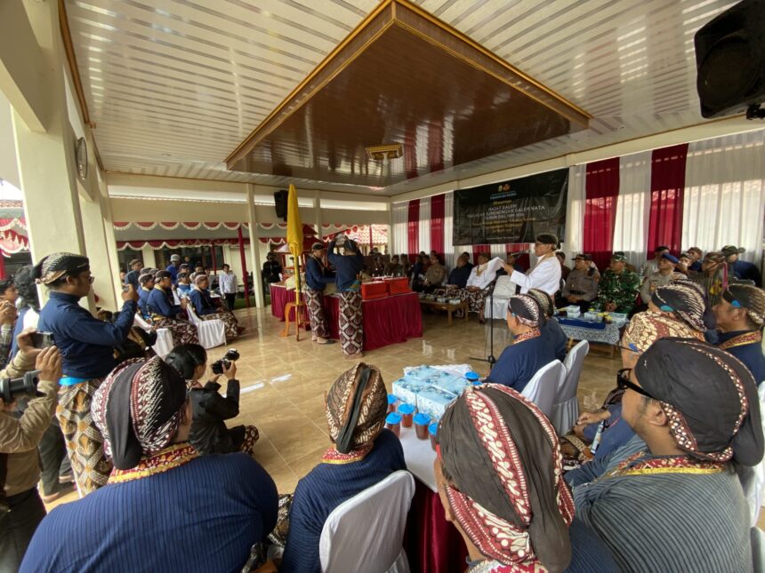 Ceremonial gathering in a hall with people in traditional Indonesian attire seated around a decorated stage and banner in the background