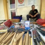 Smiling man sits on a chair outside a shop, surrounded by long wooden sticks and tools laid out on a table. A cardboard box and red baskets sit nearby.