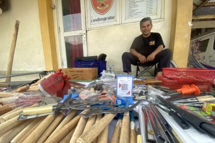 Smiling man sits on a chair outside a shop, surrounded by long wooden sticks and tools laid out on a table. A cardboard box and red baskets sit nearby.