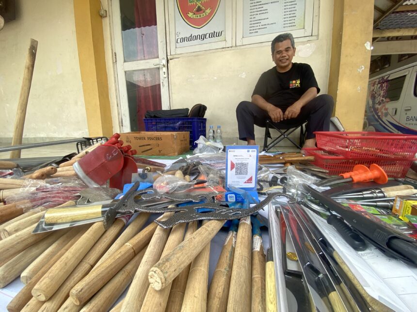 Smiling man sits on a chair outside a shop, surrounded by long wooden sticks and tools laid out on a table. A cardboard box and red baskets sit nearby.
