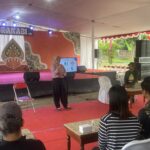 Speaker in a pink shirt presenting on a decorated stage under a white tent to an audience seated on red carpeted area