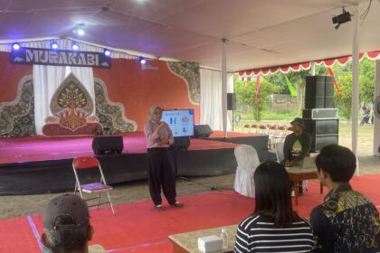 Speaker in a pink shirt presenting on a decorated stage under a white tent to an audience seated on red carpeted area