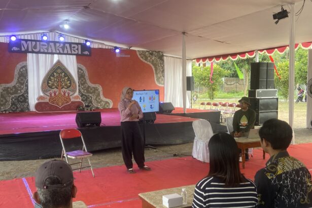 Speaker in a pink shirt presenting on a decorated stage under a white tent to an audience seated on red carpeted area
