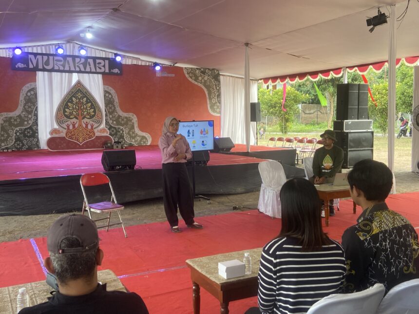 Speaker in a pink shirt presenting on a decorated stage under a white tent to an audience seated on red carpeted area