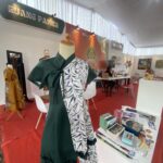 Mannequin in a green dress with a tropical-patterned wrap at an exhibition booth; tables with books and artworks visible in the background.