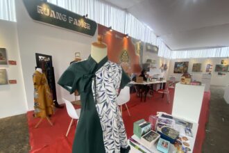Mannequin in a green dress with a tropical-patterned wrap at an exhibition booth; tables with books and artworks visible in the background.
