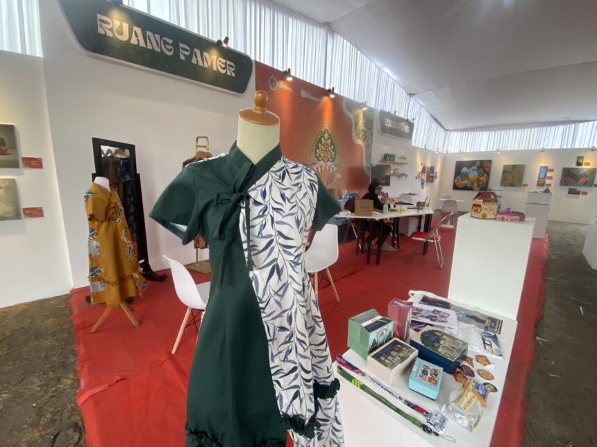 Mannequin in a green dress with a tropical-patterned wrap at an exhibition booth; tables with books and artworks visible in the background.