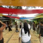 Group of people standing and raising hands under colorful fabric canopies in a courtyard.
