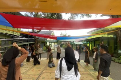 Group of people standing and raising hands under colorful fabric canopies in a courtyard.
